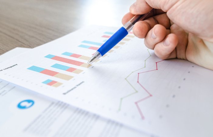 A close-up of a hand with a pen analyzing data on colorful bar and line charts on paper.