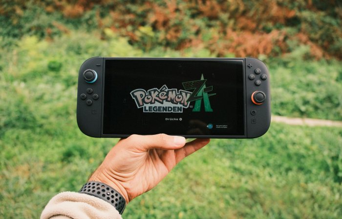 A hand holding a handheld gaming console displaying the Pokémon Legends game screen outdoors.